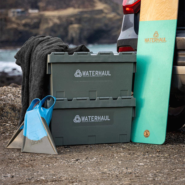 Green storage bins with 'Waterhaul' branding, a bellyboard, a towel, and swim fins against a vehicle on a beach.