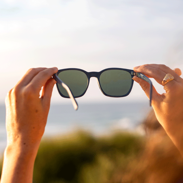 Navy sunglasses held against the sky in a coastal setting at sunset