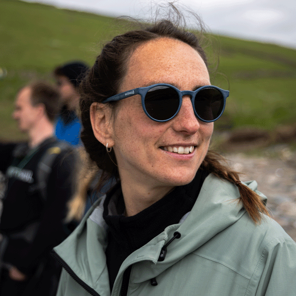 Person wearing sunglasses and a light jacket outdoors with green hills in the background
