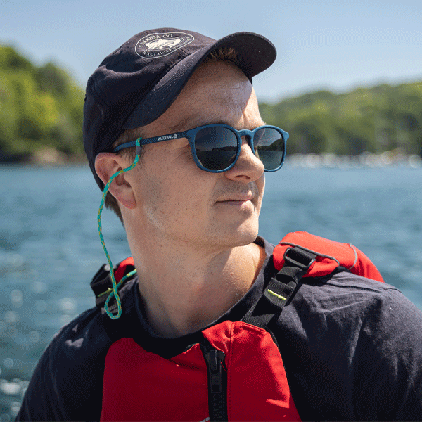 Man wearing a black cap, sunglasses, and red life jacket by a body of water.