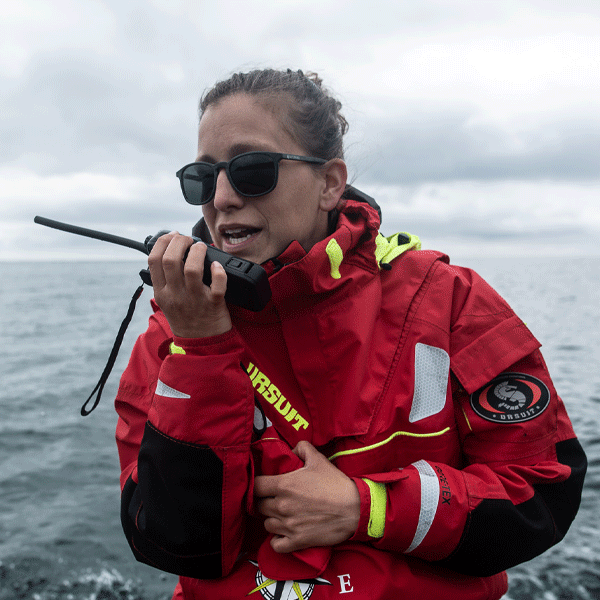 Person wearing sunglasses in a red life jacket using a radio near water