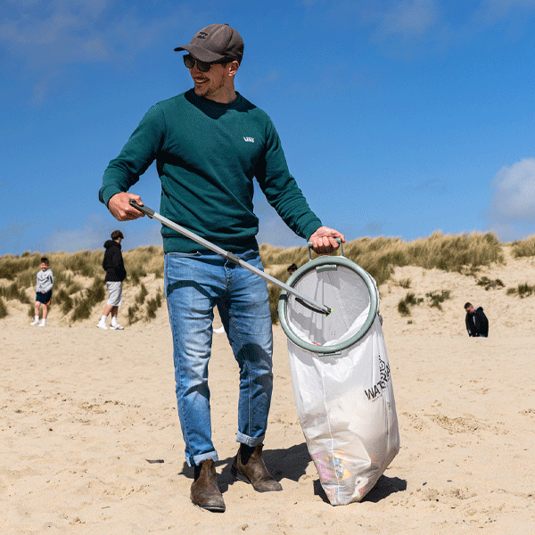 Man on a beach holding a litter picker and trash bag, with a clear blue sky and dunes in the background.
