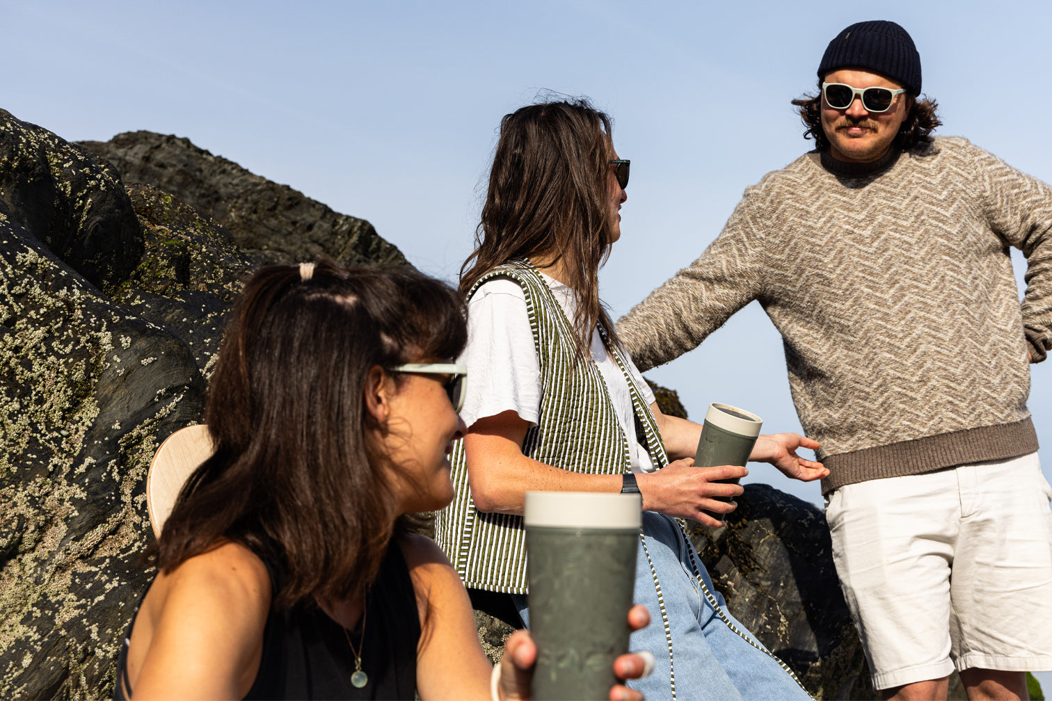 Three people standing outdoors with a clear blue sky, two people holding a coffee cup.