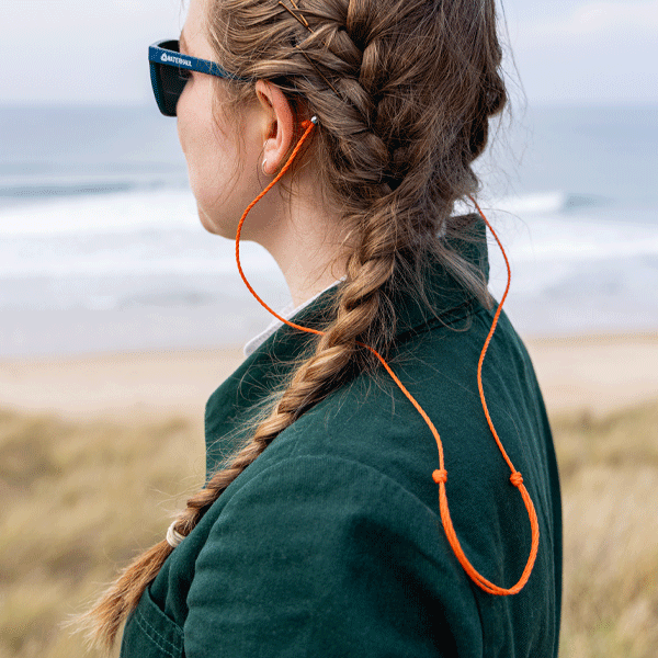 Person with braided hair wearing sunglasses and green jacket, standing by a body of water.