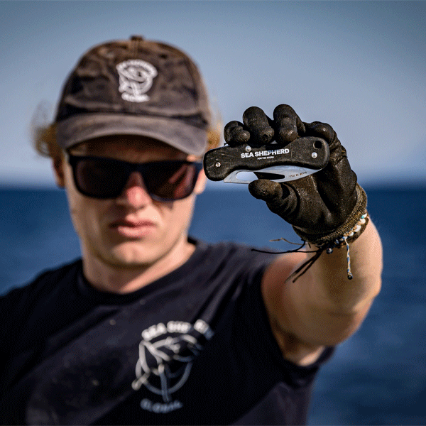 Person wearing a cap and sunglasses, holding a Sea Shepherd tool against a blue background
